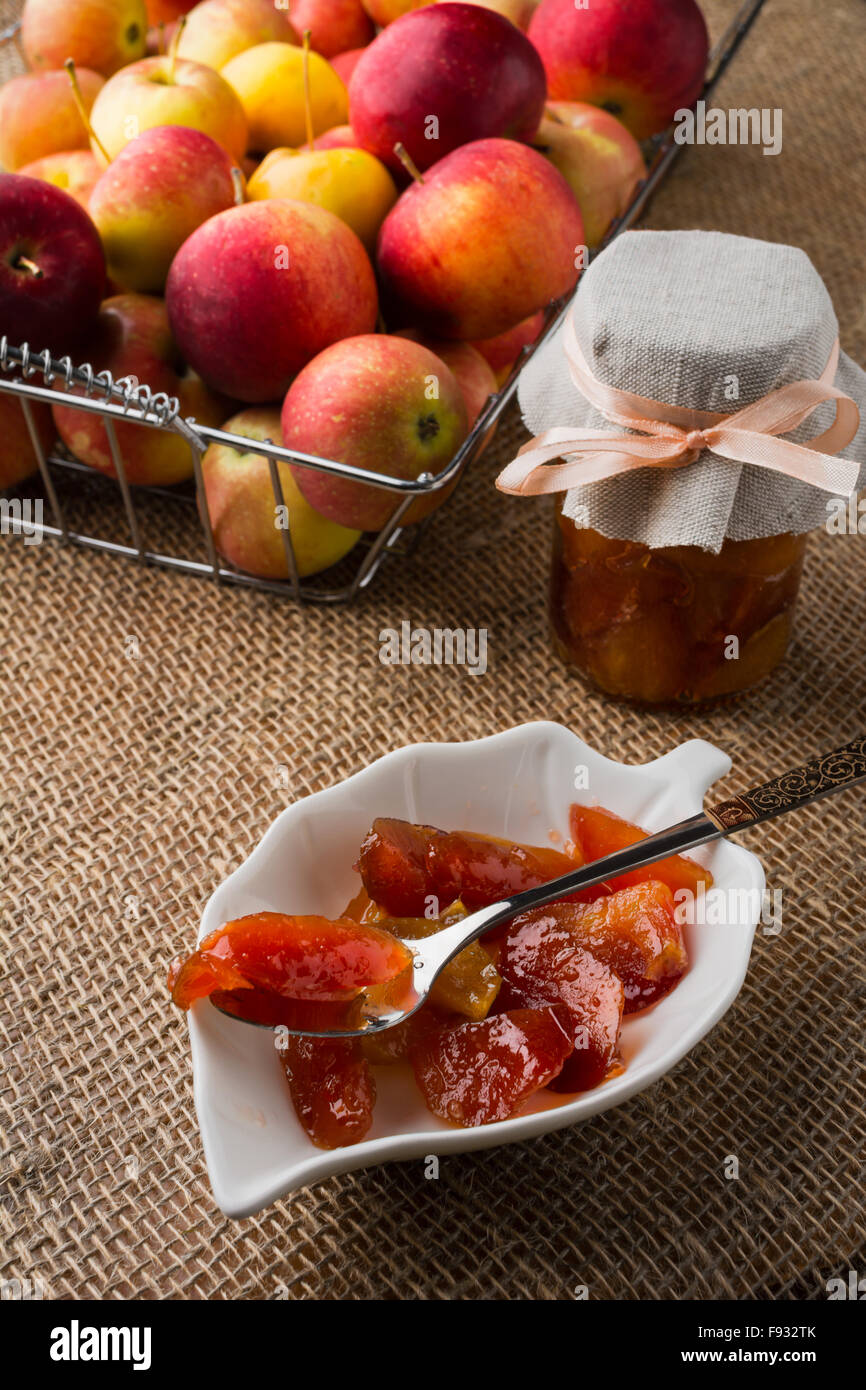 In casa delle fette di marmellata di mele su bianco piattino, cucchiaio, un vasetto di marmellata di arance e mele in filo metallico cesto su una tela coperti da tavola, Foto Stock