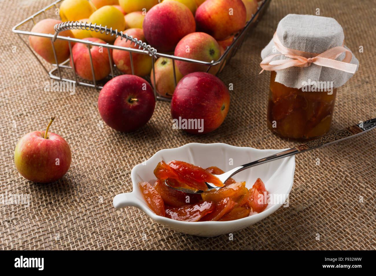 In casa la marmellata di mele su bianco piattino, cucchiaio, un vasetto di marmellata di arance e mele in filo metallico cesto su un tavolo ricoperto con tela. Sele Foto Stock