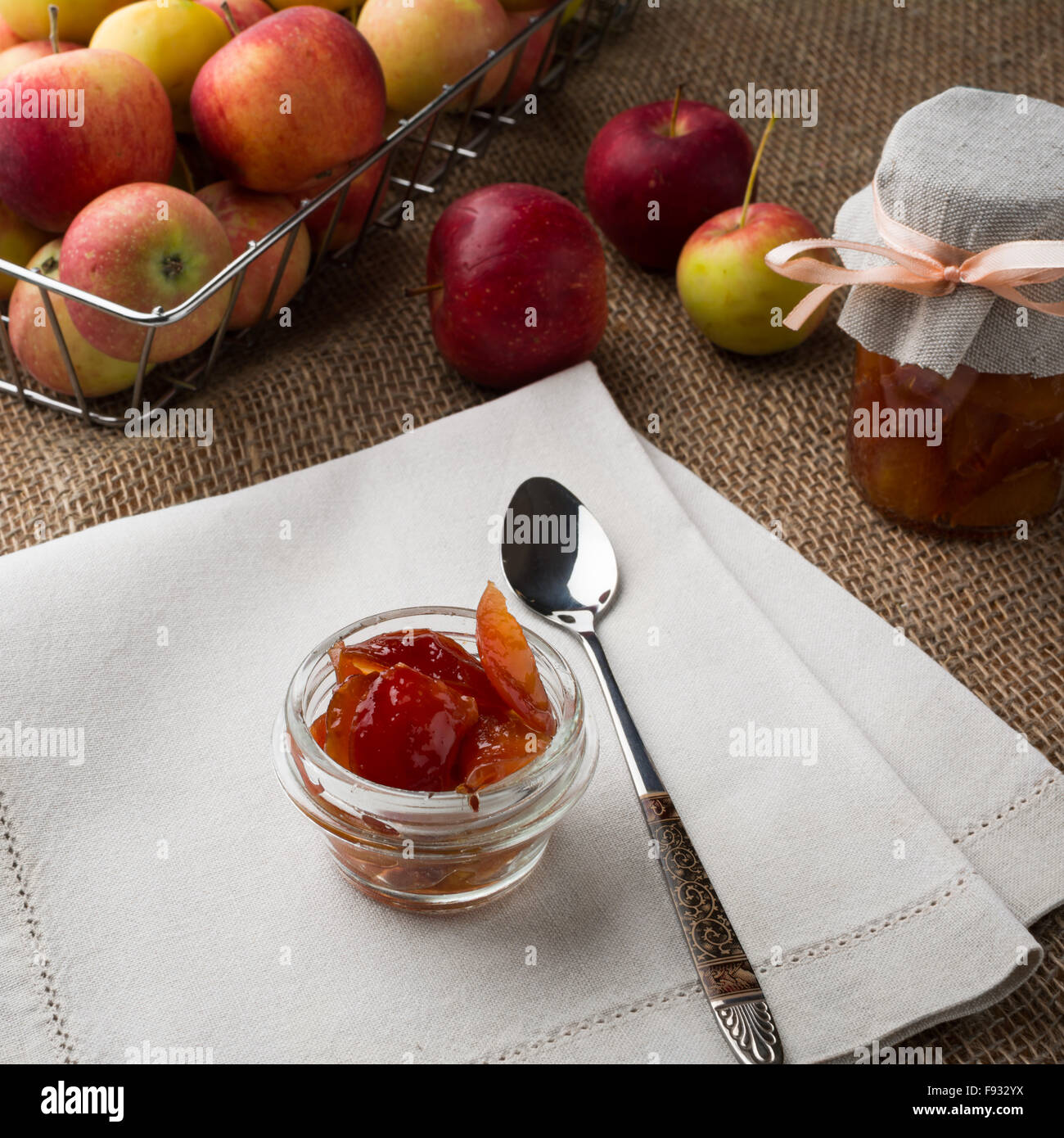 Il vasetto di vetro di marmellata di mele, cucchiaio, un vasetto di marmellata di arance, biancheria igienico e mele in filo metallico cesto su una tela coperti da tavola, square. Foto Stock