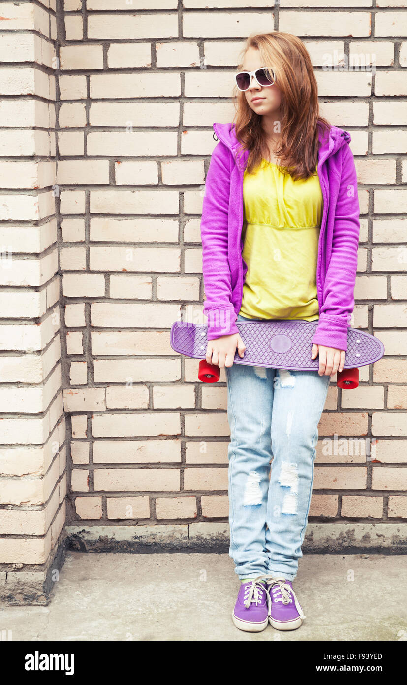 La bionda ragazza adolescente in occhiali da sole e colorato i vestiti  sportivi skateboard tiene vicino dal grigio urbano parete di mattoni Foto  stock - Alamy