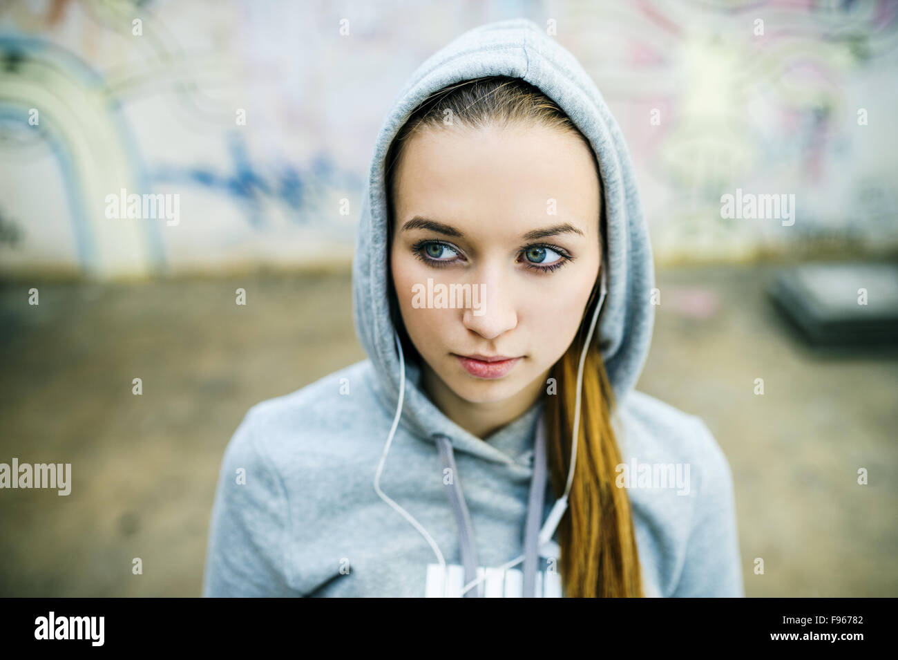 Grave ragazza adolescente con cappa in ascolto di musica dalle cuffie in piedi su sfondo del muro di graffiti Foto Stock