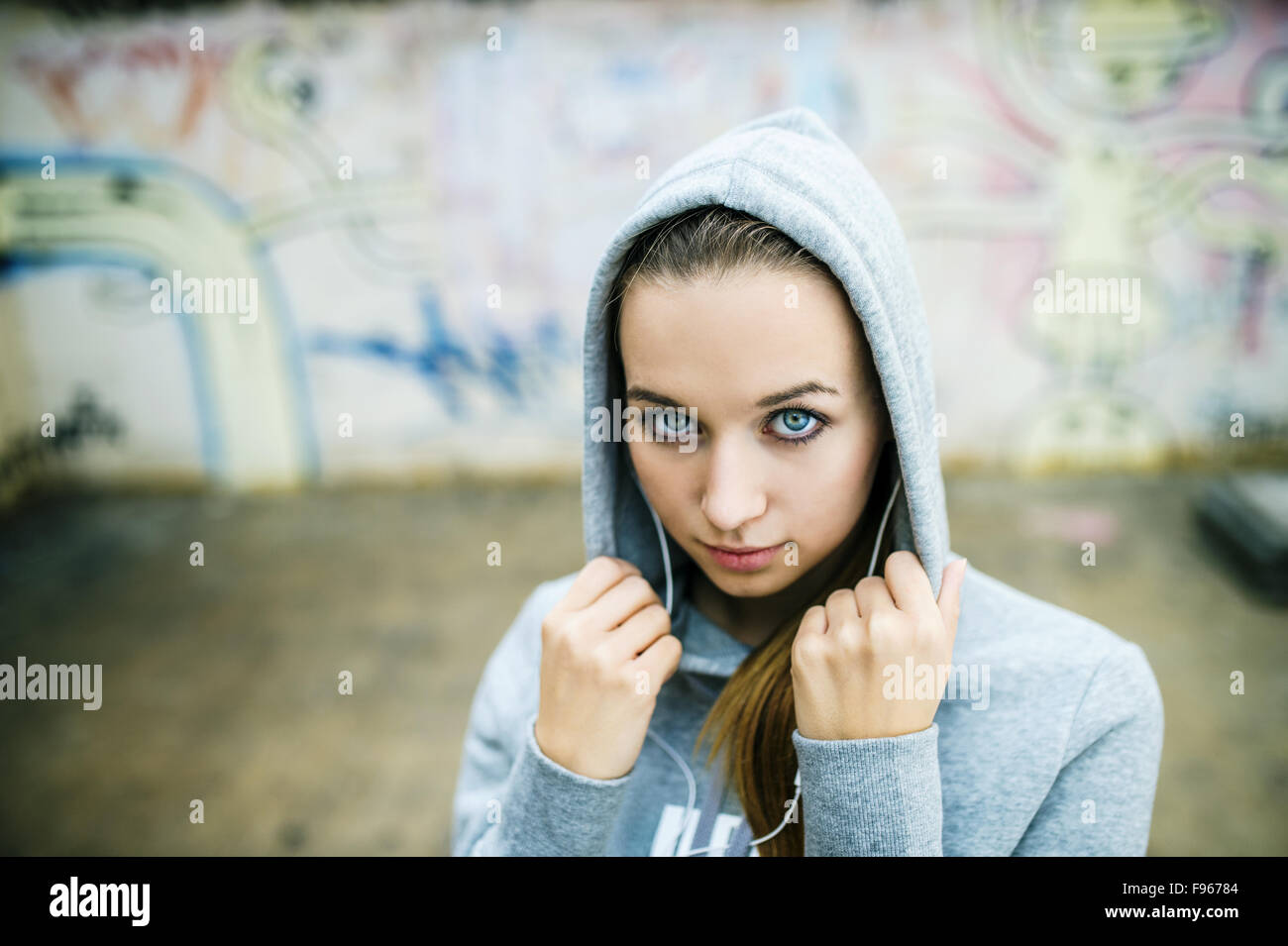 Grave ragazza adolescente con cappa in ascolto di musica dalle cuffie in piedi su sfondo del muro di graffiti Foto Stock