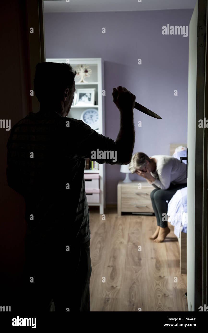 Donna matura siiting sul letto è spaventato da uomo con coltello. La donna è vittima di violenza domestica e abusi. Foto Stock