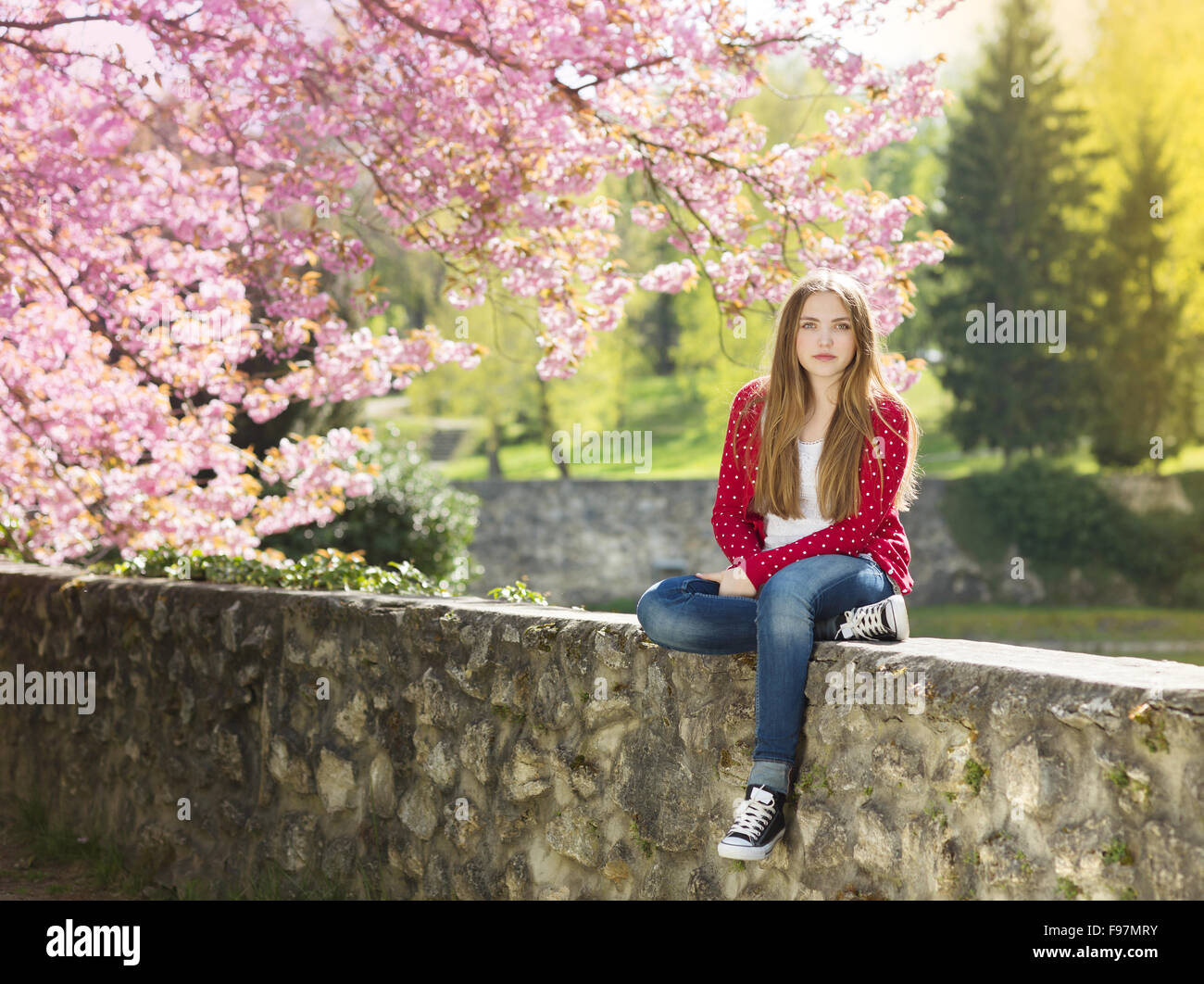 Bella ragazza in rosso cardigan seduto su una parete nella primavera del giardino con alberi in fiore Foto Stock