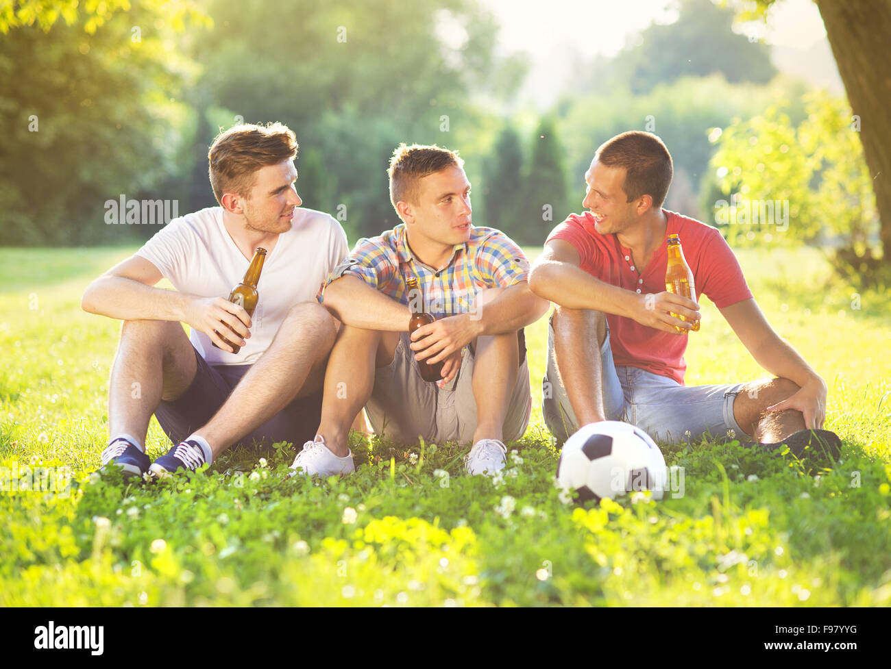 Tre amici felice trascorrere del tempo libero insieme nel parco seduto su erba, bere birra e chat Foto Stock