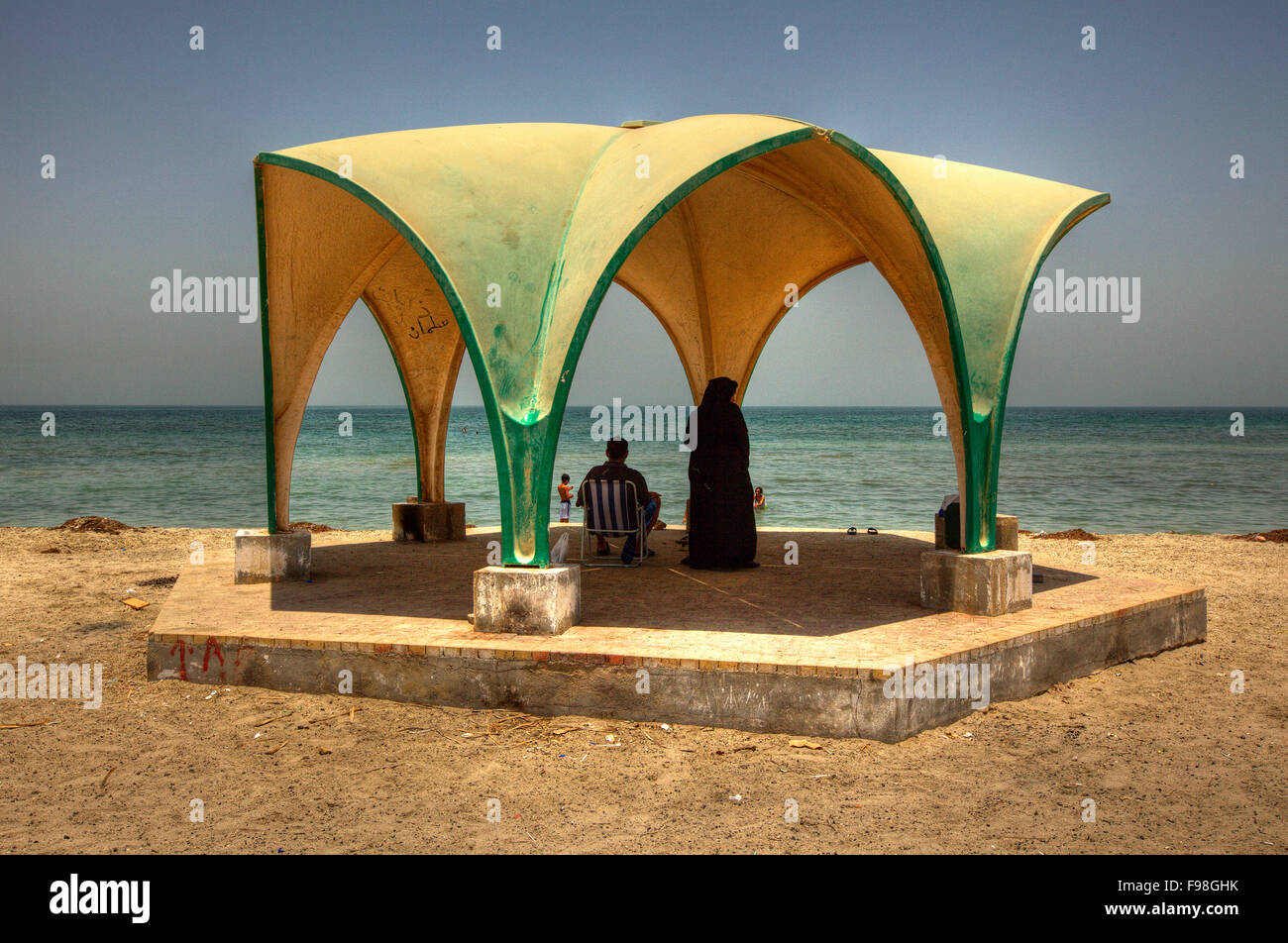 Un paio di persone locali in ombra a guardare i bambini in primo piano la riproduzione nel mare di Al Jazayer Beach, isola di Bahrain. Foto Stock