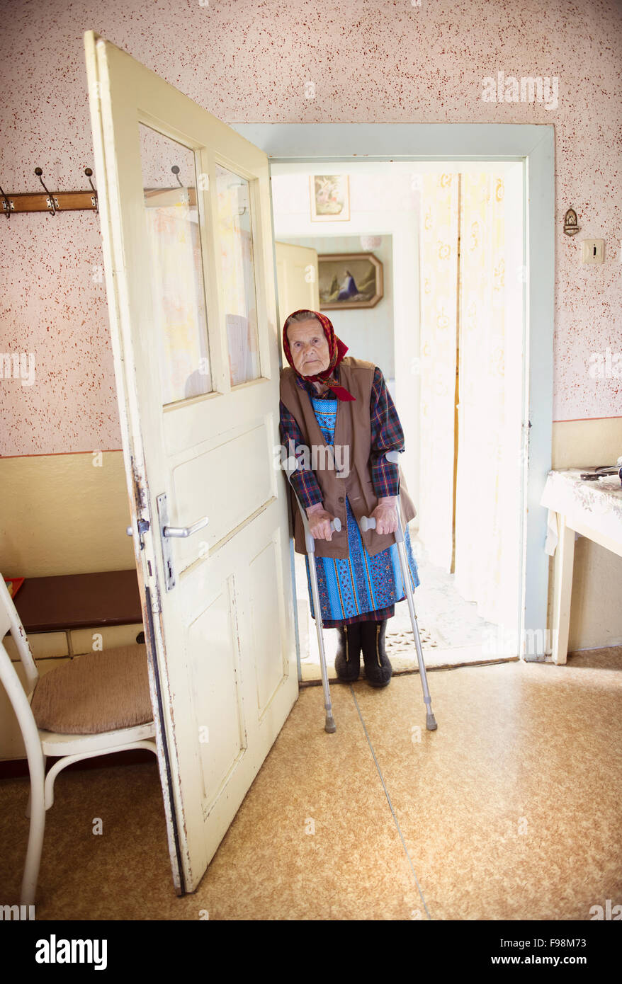 Molto vecchia donna è in piedi per la porta con le stampelle al suo stile country house Foto Stock