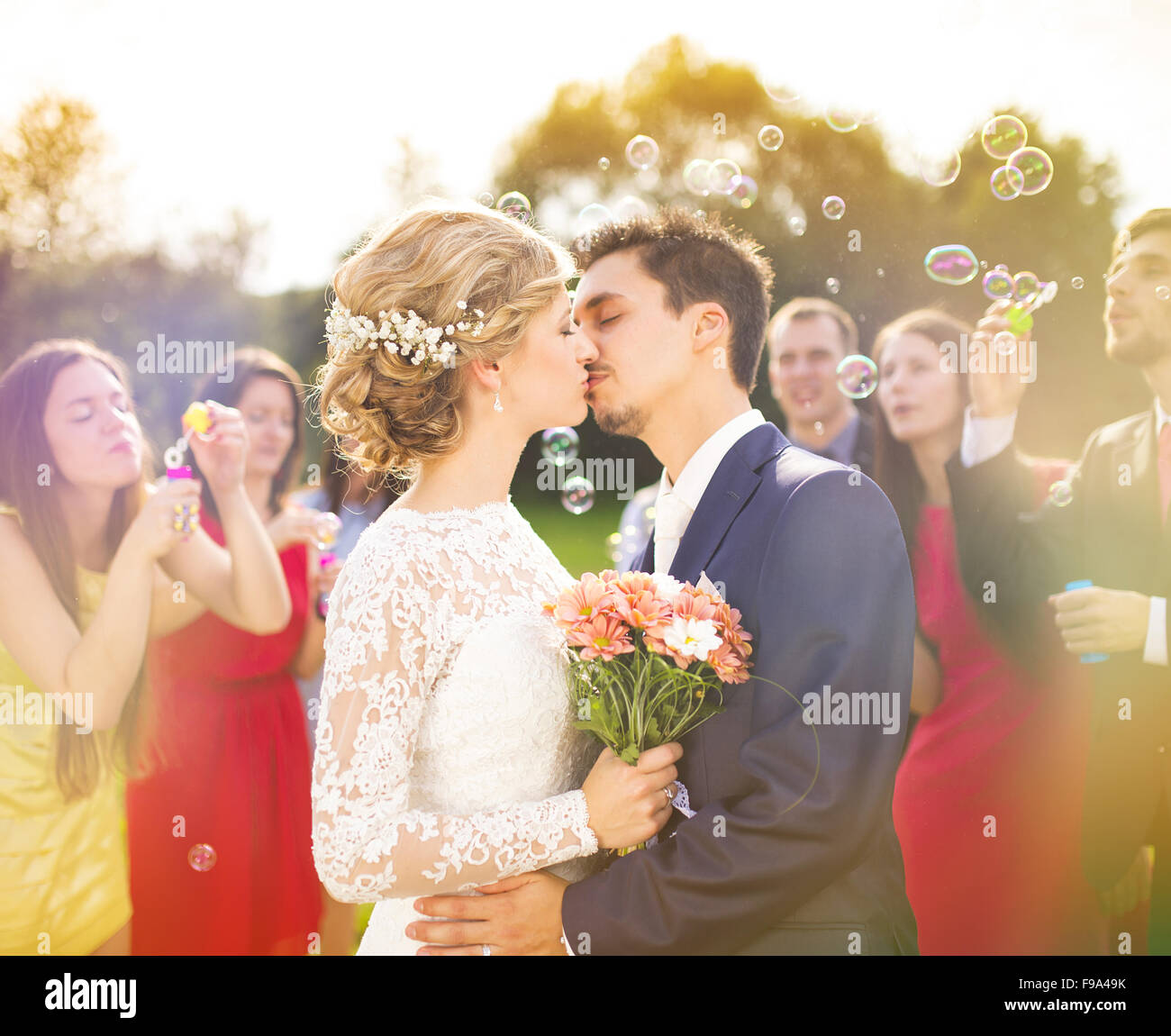 I giovani sposi novelli baciare e godendo di momento romantico insieme al ricevimento di nozze al di fuori, ospiti di nozze nel soffiaggio di sfondo Foto Stock