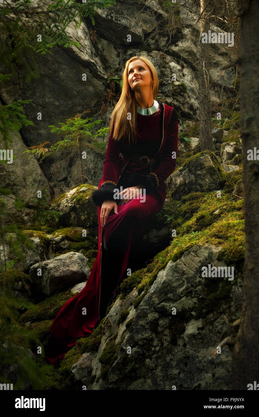 Una donna bellissima fata con lunghi capelli biondi in un abito storico è seduta amids moos rocce coperte in incantevole paesaggio forestral Foto Stock