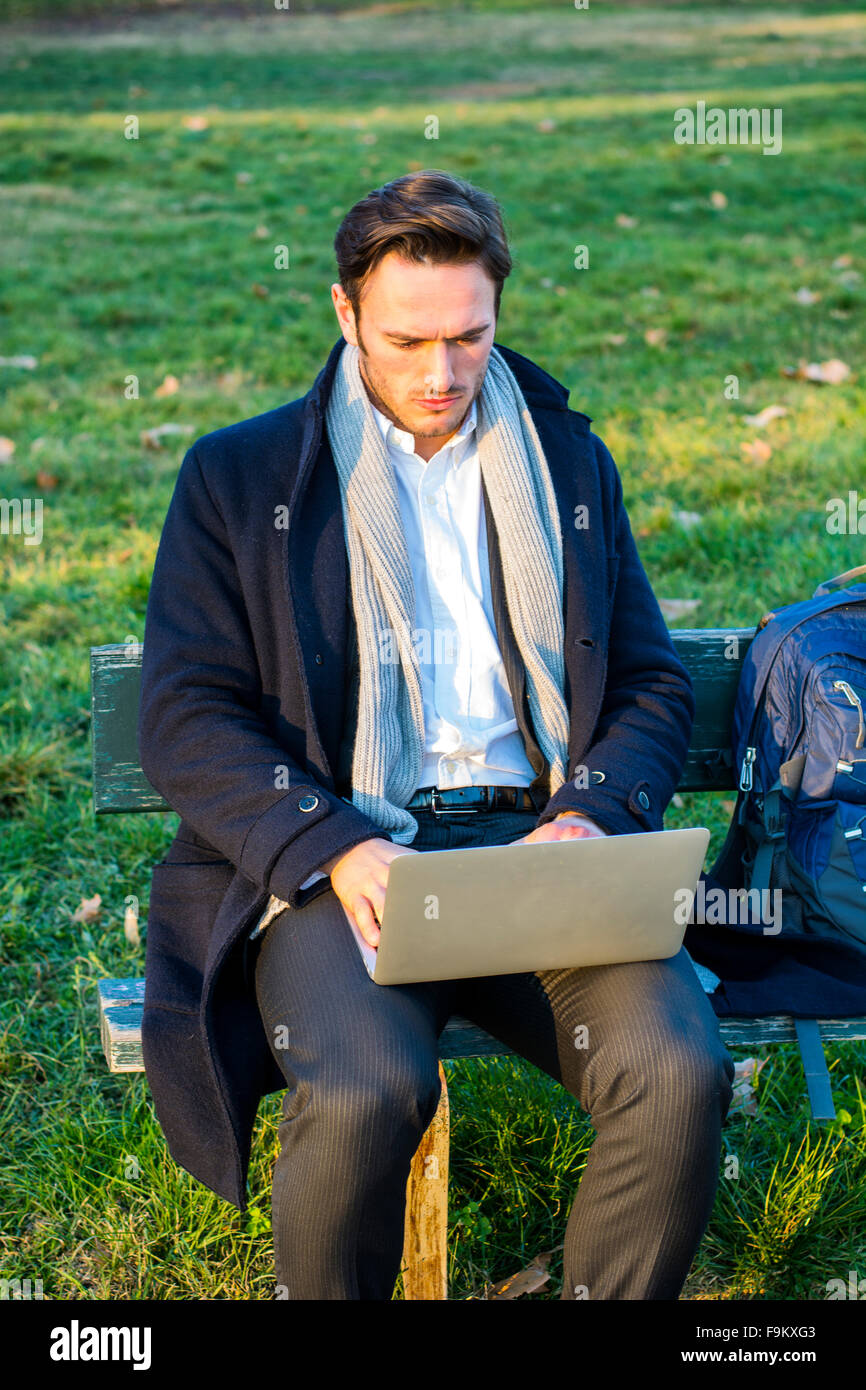 Bello ed elegante imprenditore seduto su una panca in legno per lavorare in esterno in un parco urbano di digitare le informazioni sul suo computer portatile comp Foto Stock