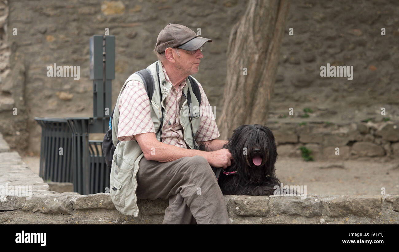 Un vecchio uomo che indossa berretto seduto su una parete con la sua black shaggy pelose bouvier de flandres cane Foto Stock