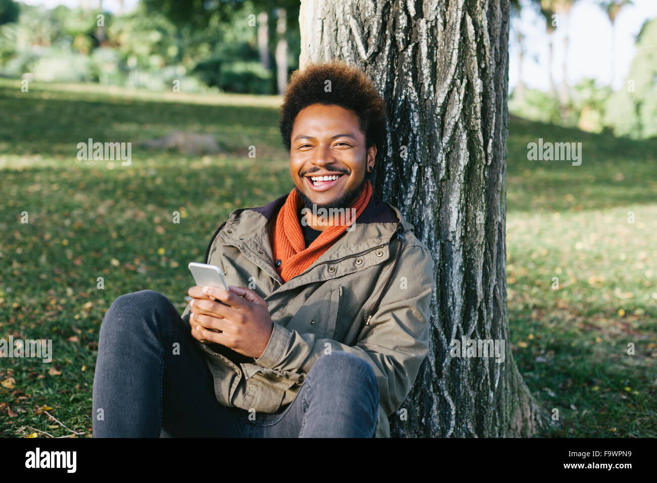 Ritratto di giovane sorridente uomo con lo smartphone appoggiato contro il tronco di albero Foto Stock