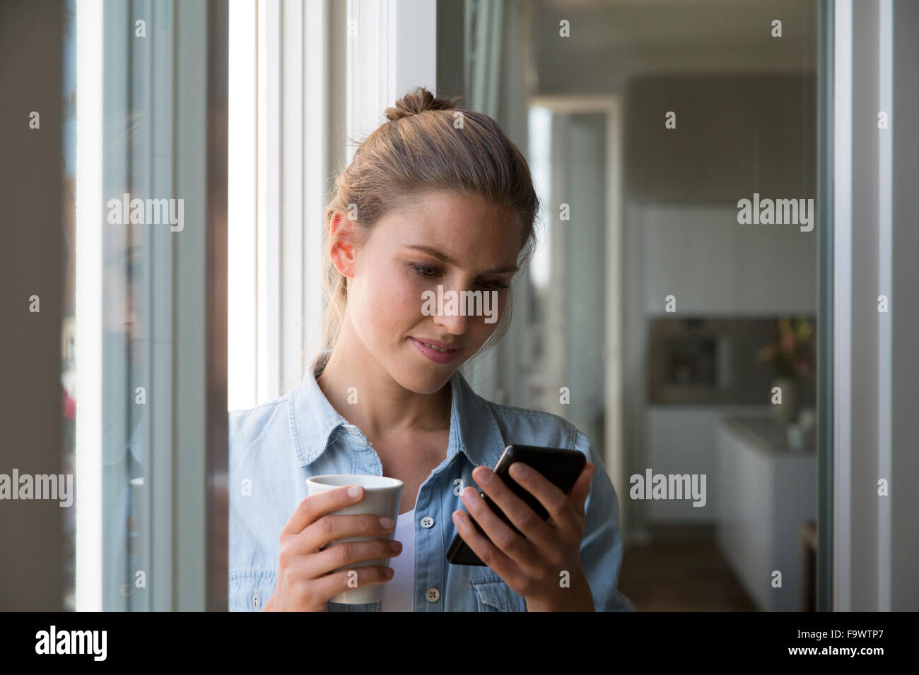 Giovane donna in piedi presso la finestra con smart phone e tazza da caffè Foto Stock