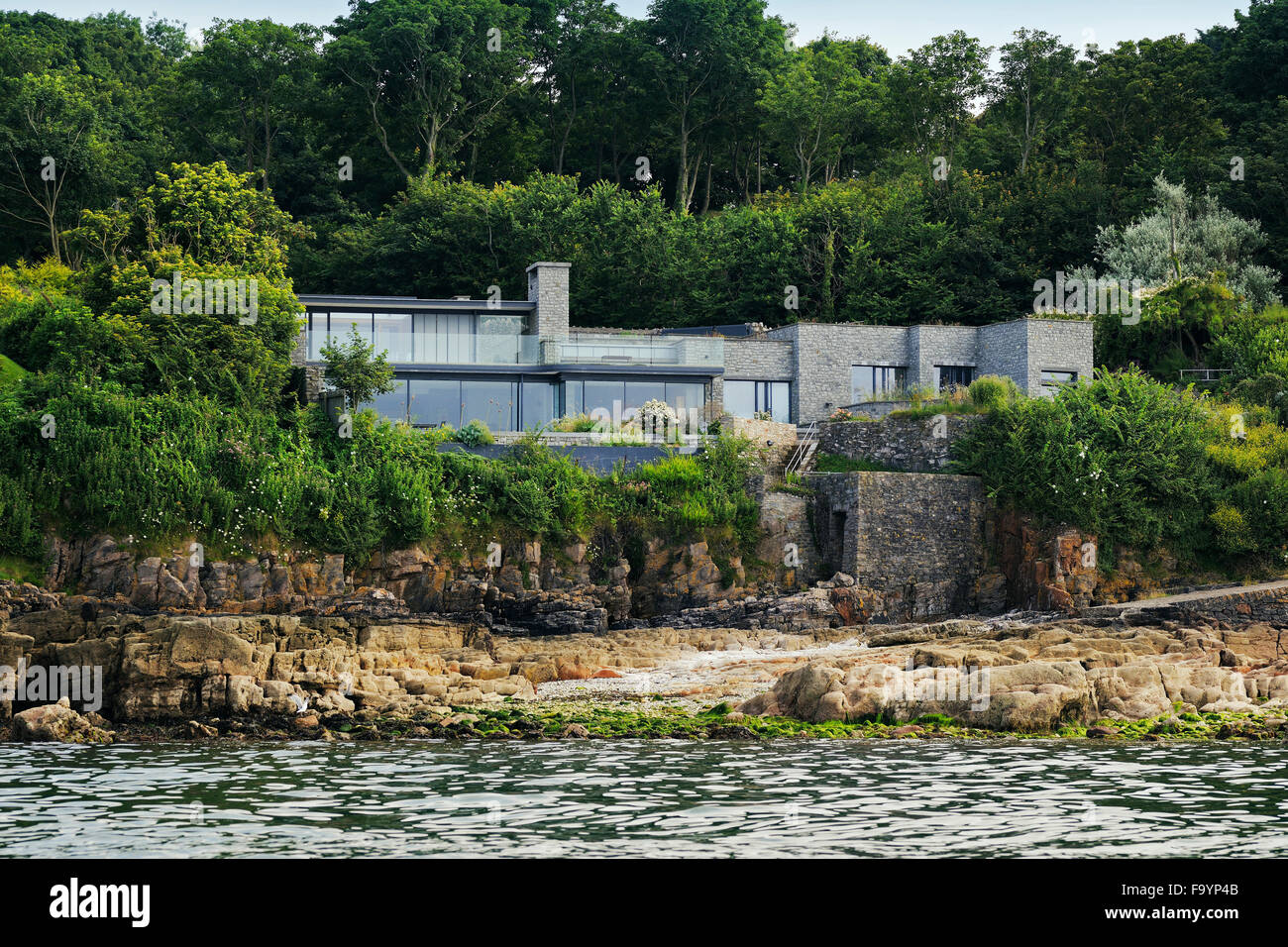 Vista dall'acqua di un seafont clifftop casa moderna dal patio, guardando alla principale spazio abitativo. Grandi porte scorrevoli in vetro aperto. Il pavimento e il soffitto di pannelli di vetro. Un basso edificio di profilo in una posizione sul mare. Foto Stock