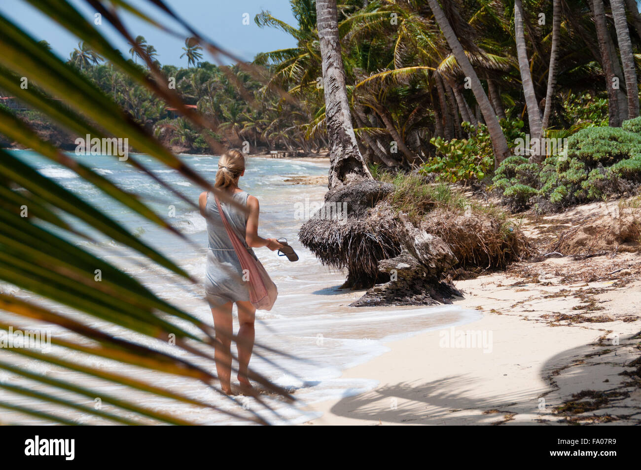 Bella Donna bionda con borsa rosa camminare a piedi scalzi lungo la solitaria dei caraibi spiaggia di sabbia bianca e palme di cocco dietro lascia sulla piccola isola di mais Foto Stock