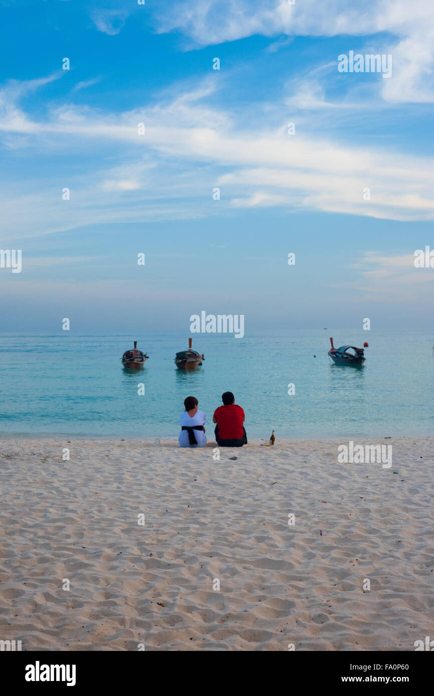 Un turista giovane seduto sulla sabbia bianca, la condivisione di una tranquilla vista oceano momento sulla spiaggia davanti ad un mare calmo e cielo blu chiaro Foto Stock