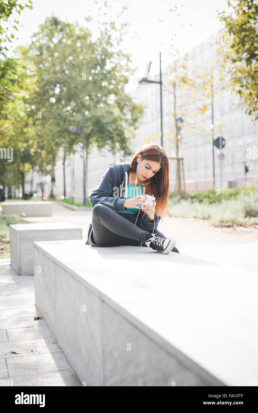 Giovane bello caucasica capelli rossicci donna seduta su una piccola parete, ascoltando musica con le cuffie e lo smartphone maniglia - musica, relax, il concetto di tecnologia Foto Stock