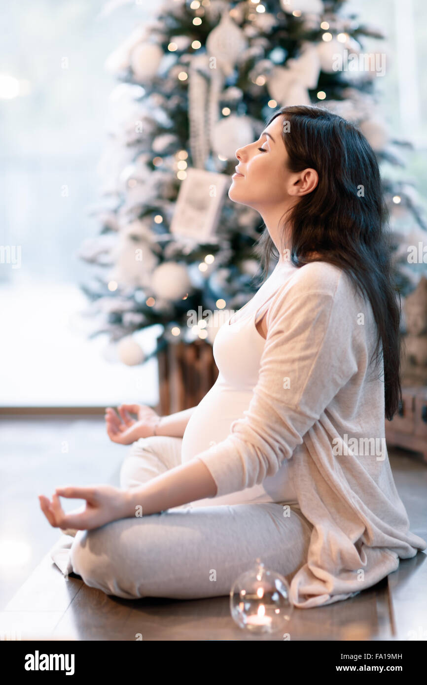 Donna incinta meditando a casa vicino al bellissimo albero di Natale decorato, seduta in lotus posano con gli occhi chiusi Foto Stock