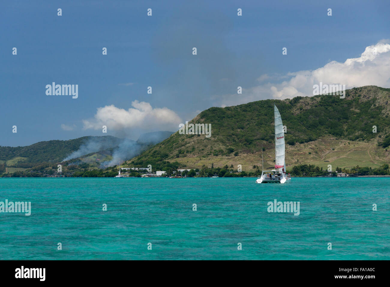Un catamarano turistico off sulla costa est di Mauritius, con la masterizzazione di canna da zucchero sulla riva Foto Stock