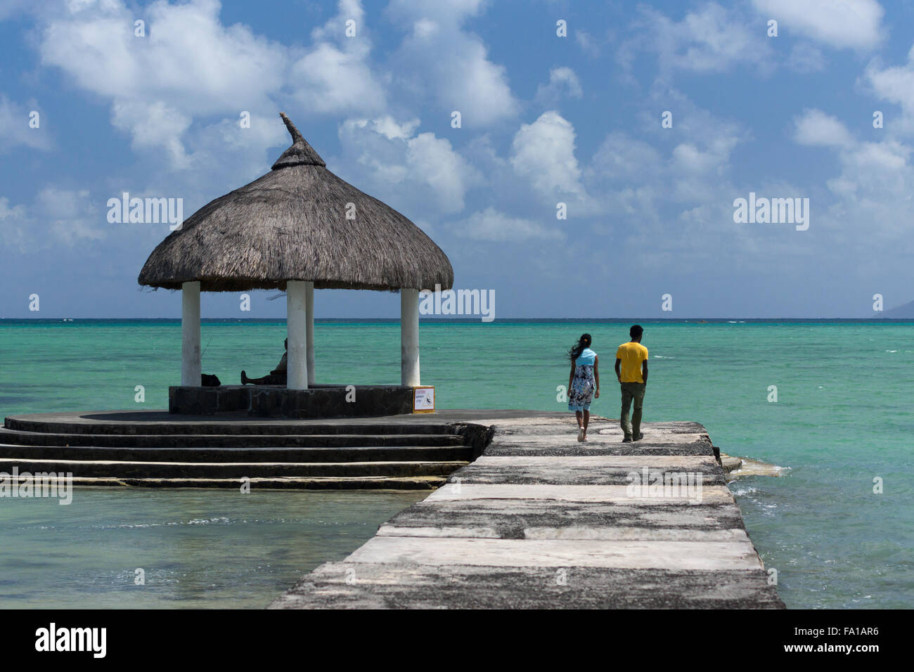 Un paio di mauriziani che camminano lungo un molo verso l'Oceano Indiano con una capanna sulla spiaggia alla fine del molo. Grand Gaube, Mauritius Foto Stock