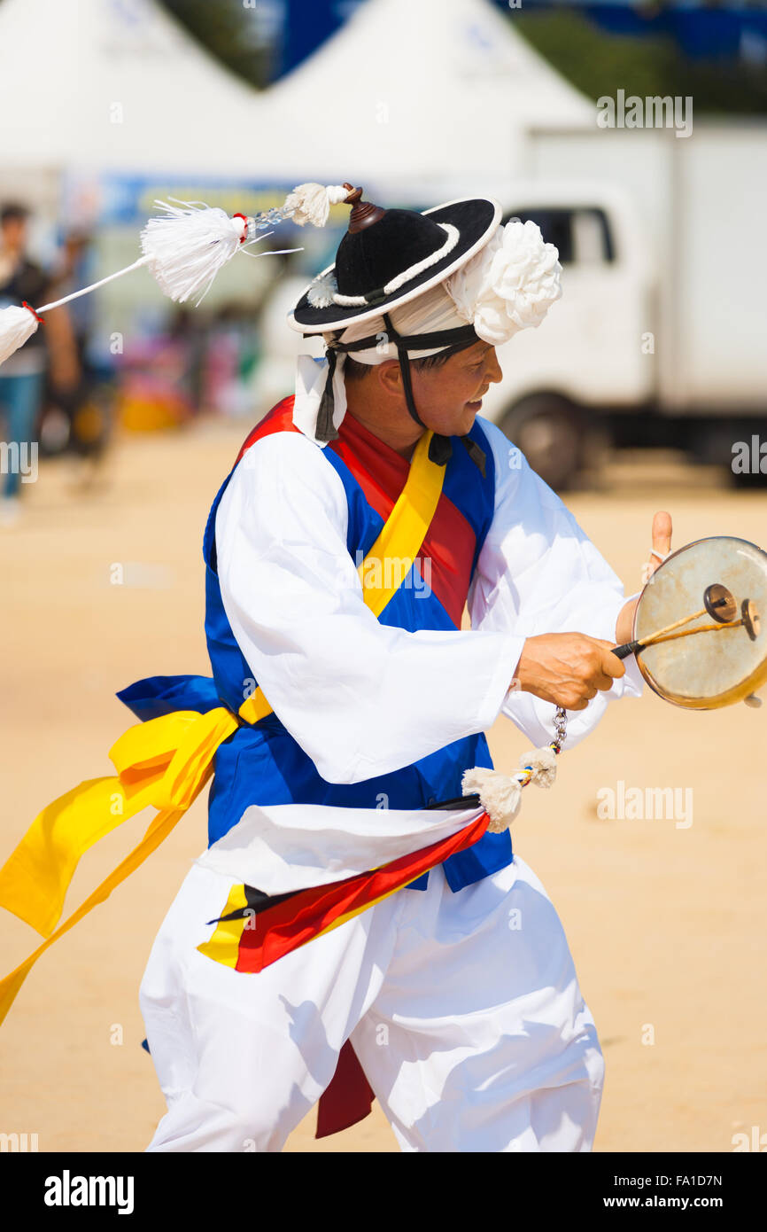 Un coreano uomo vestito di bianco vestiti tradizionali giubbotto blu dancing e drumming durante la filatura il suo cappello tassle al festival locale Foto Stock