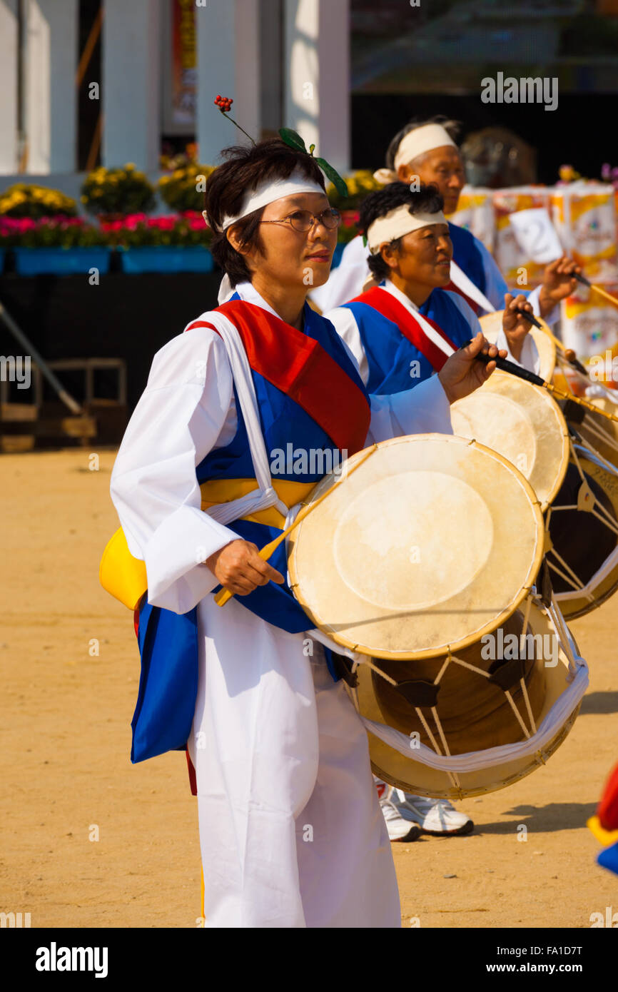 Una matura coreano donna vestita a festa, colorati abiti tradizionali battendo due facciate tamburi janggu al locale festival all'aperto Foto Stock