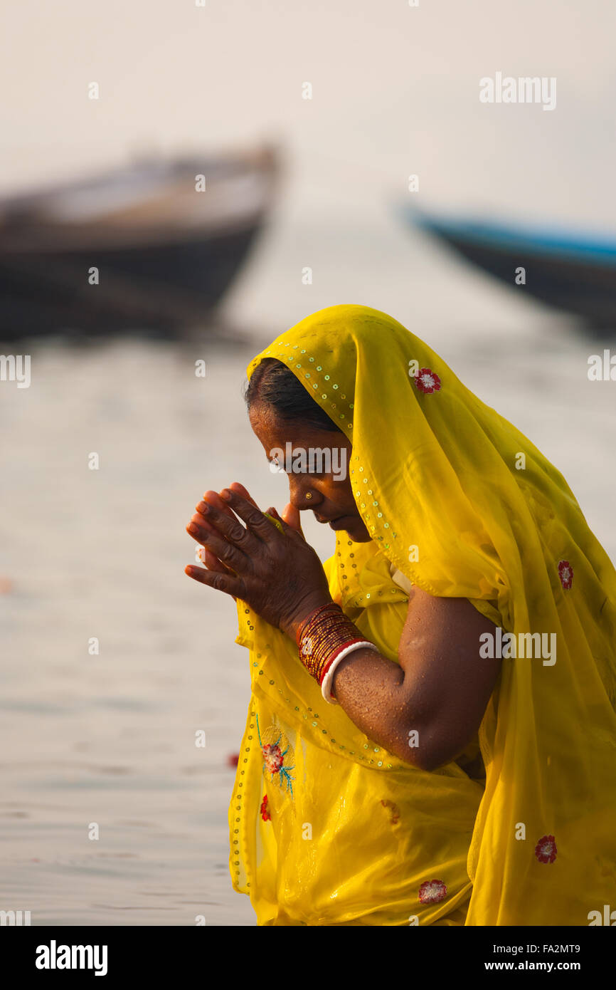 Una pia donna indù in giallo sari inarcamento, pregando e balneazione con le mani incrociate insieme nel sacro Gange fiume nel mattino Foto Stock
