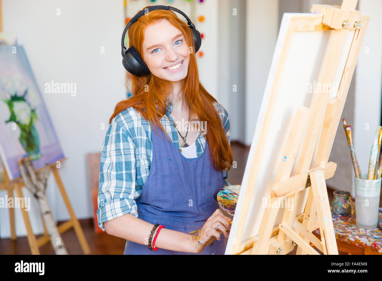 Affascinante sorridente giovane donna artista in cuffia e grembiule dipinto su tela e ascolto di musica in art studio Foto Stock