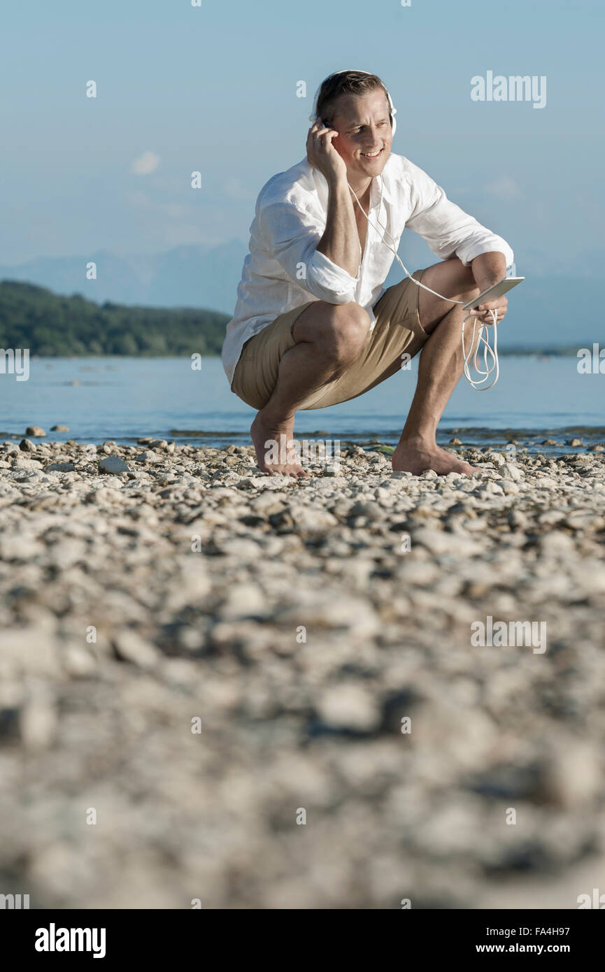 Uomo maturo seduta a Lakeside e ascolto di musica con cuffie, Baviera, Germania Foto Stock
