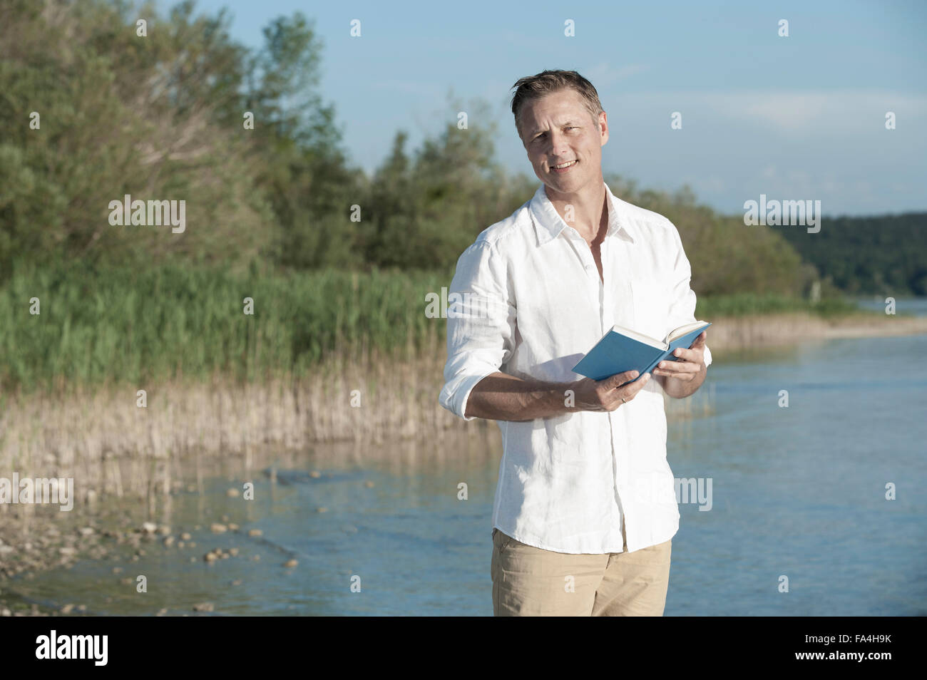 Uomo maturo la lettura del libro e sorridente a Lakeside, Baviera, Germania Foto Stock