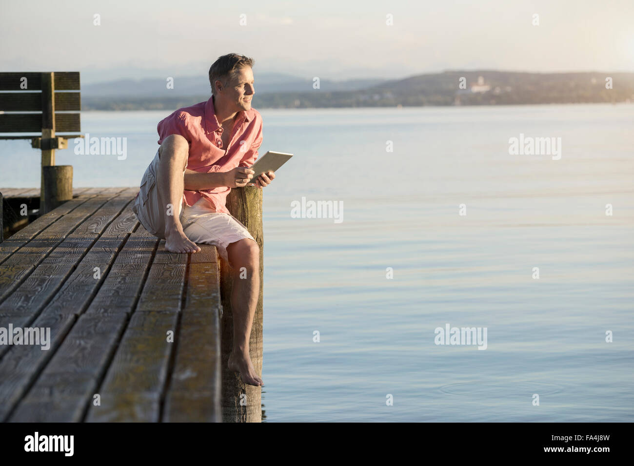 Uomo maturo con tavoletta digitale sul molo e guardando oltre il lago di Baviera, Germania Foto Stock