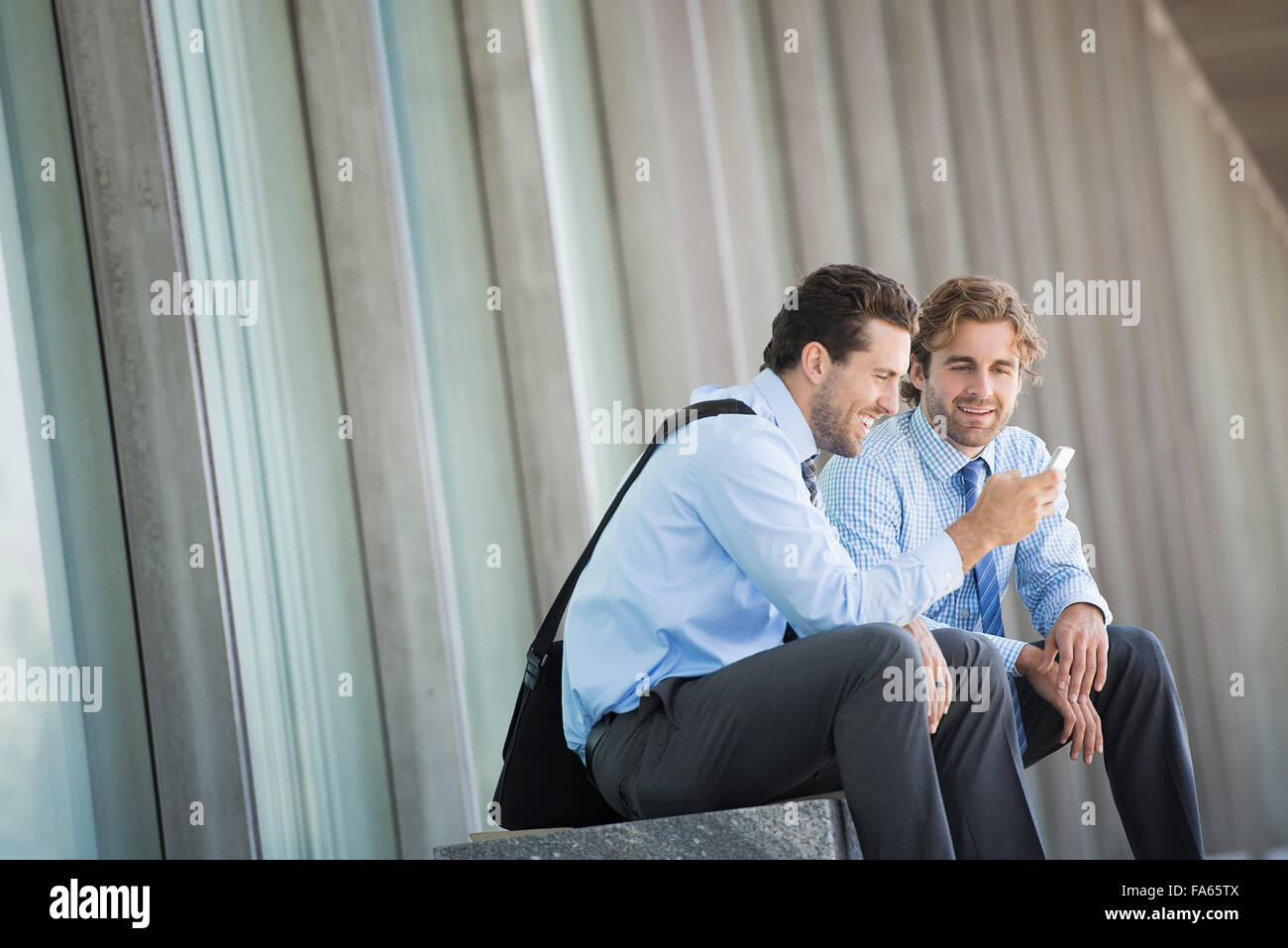 Due uomini di affari in camicie e cravatte seduta al di fuori di un edificio adibito ad uffici, controllando la loro smart phone. Foto Stock