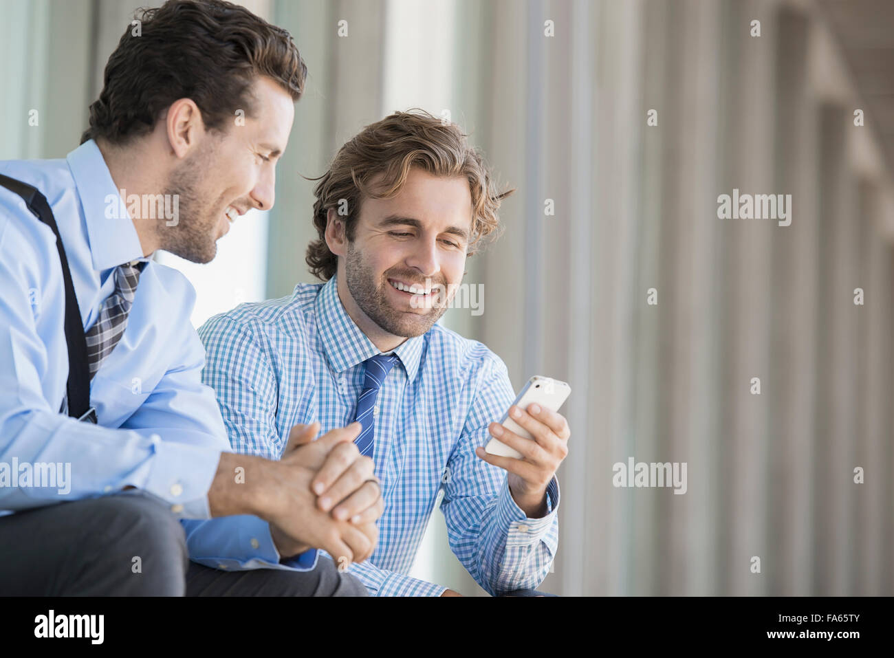 Due uomini di affari in camicie e cravatte seduta al di fuori di un edificio adibito ad uffici, controllando la loro smart phone. Foto Stock