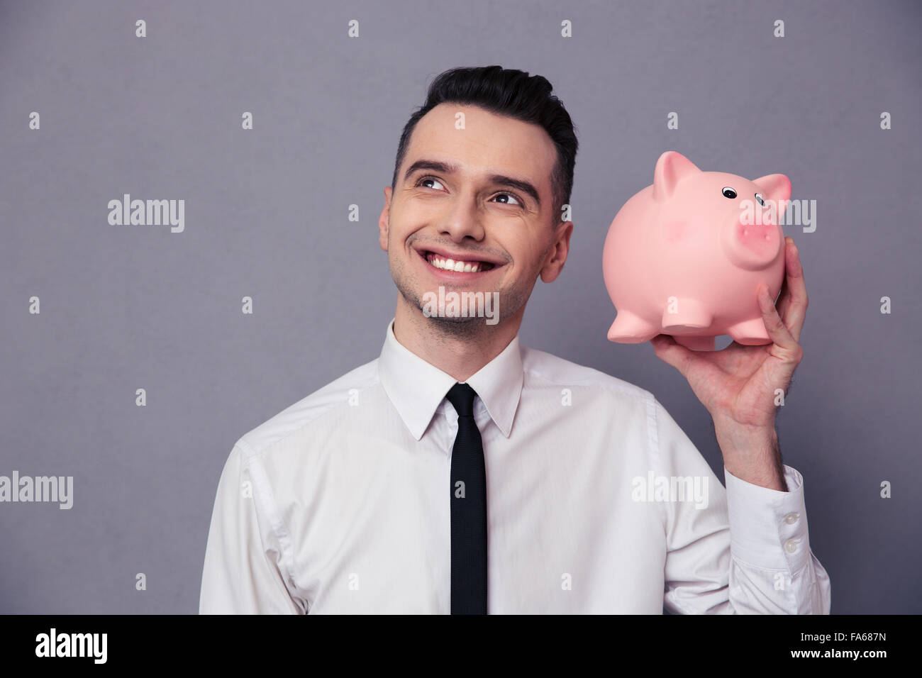 Ritratto di un happy businessman holding suino casella di denaro su sfondo grigio Foto Stock