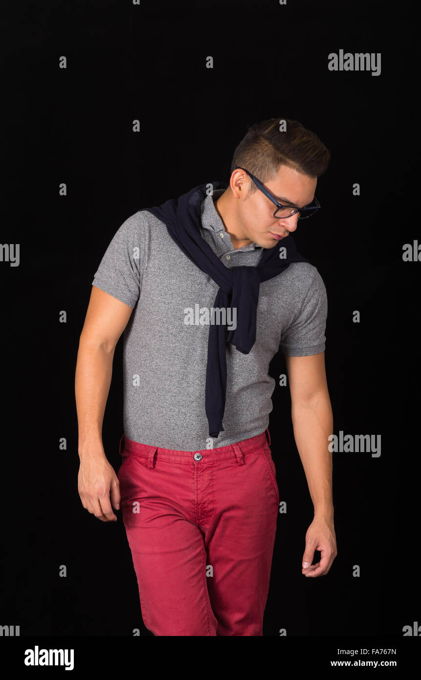 Ispanico maschio a tenuta che indossa la maglietta, maglione sulle spalle,  pantaloni rossi e bicchieri raffigurante un sofisticato stile hipster, con  lo sguardo verso il basso, sfondo nero Foto stock - Alamy