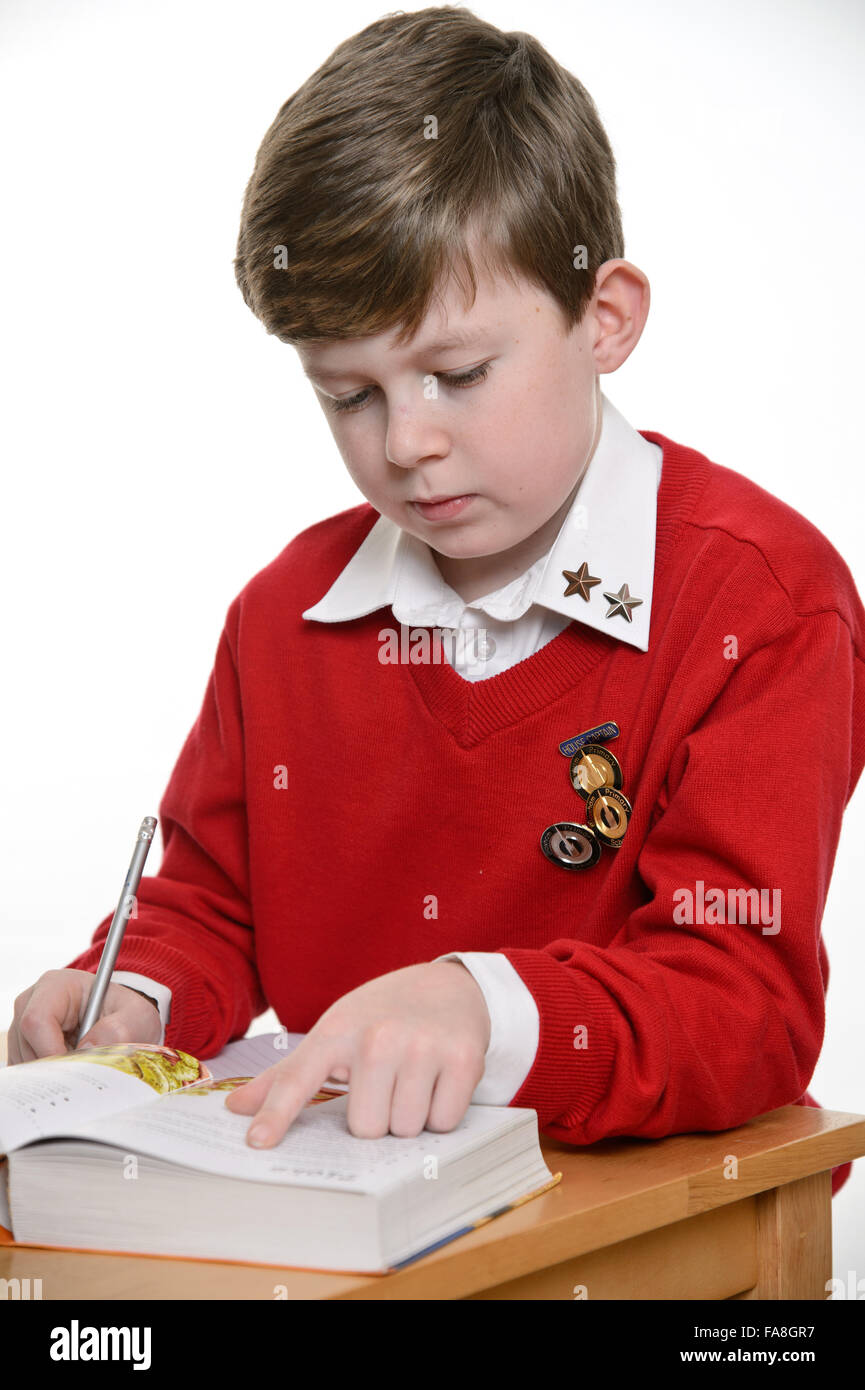 Un giovane (scuola primaria) ragazzo in un rosso uniforme scolastica maglione libri di lettura e scrittura di note per i suoi compiti. Foto Stock