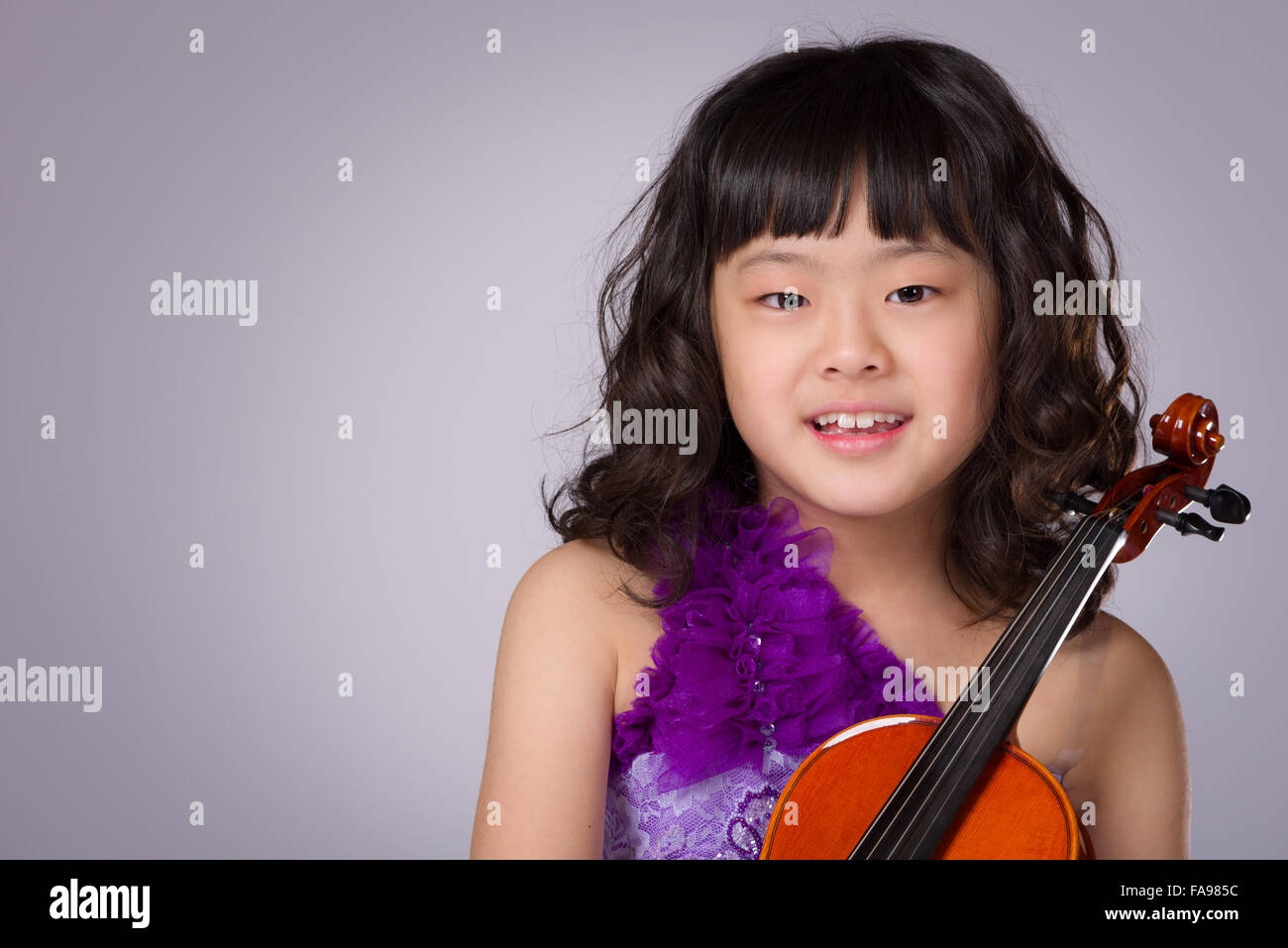 Un ritratto di un simpatico, felice e giovane giapponese ragazza in un abito viola su uno sfondo grigio con un violino. Foto Stock