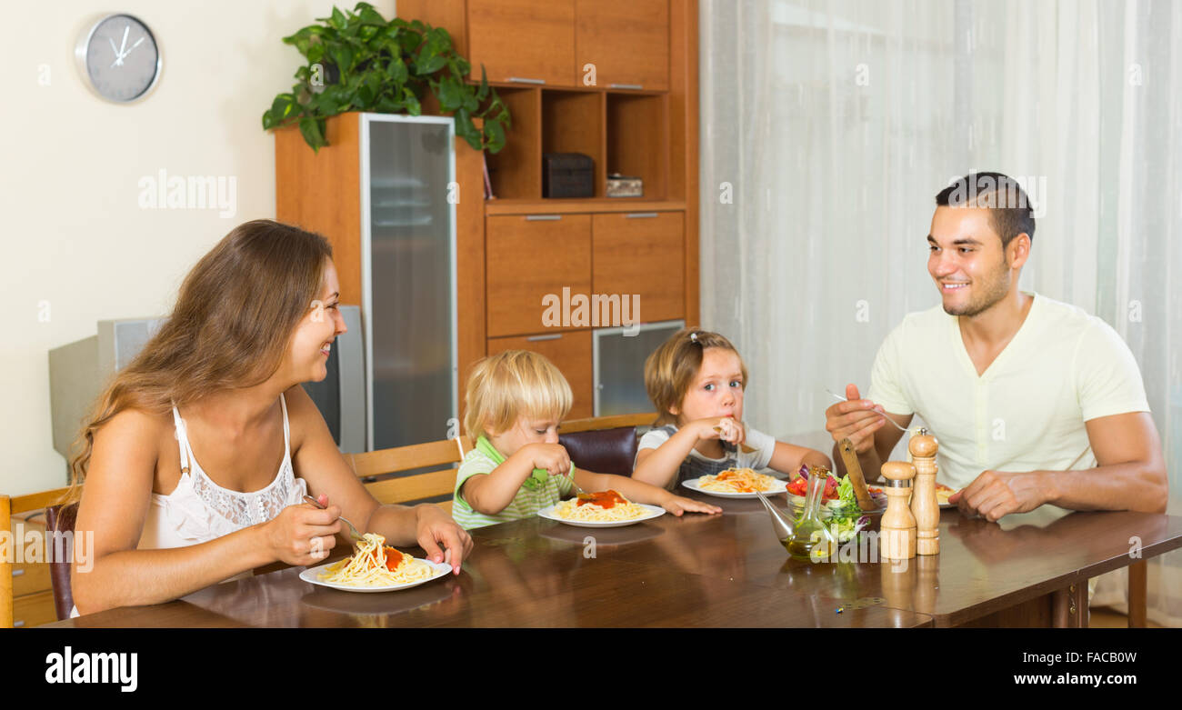 Felice famiglia giovane di quattro a pranzo con spaghetti a casa. Focus sulla donna Foto Stock