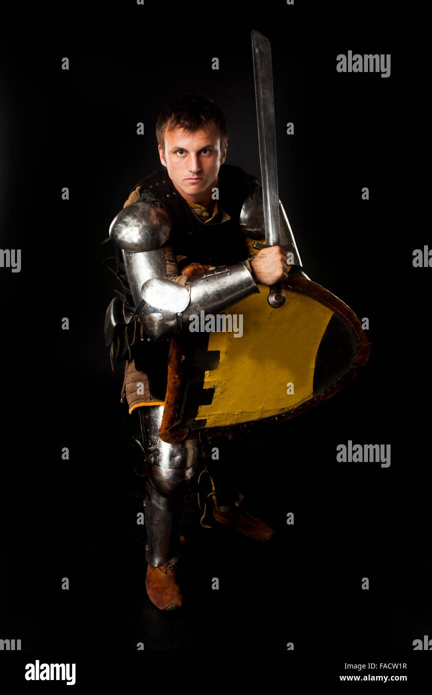 Studio shot del giovane uomo vestito da cavaliere medievale armato di spada e scudo in posizione di difesa su sfondo nero Foto Stock