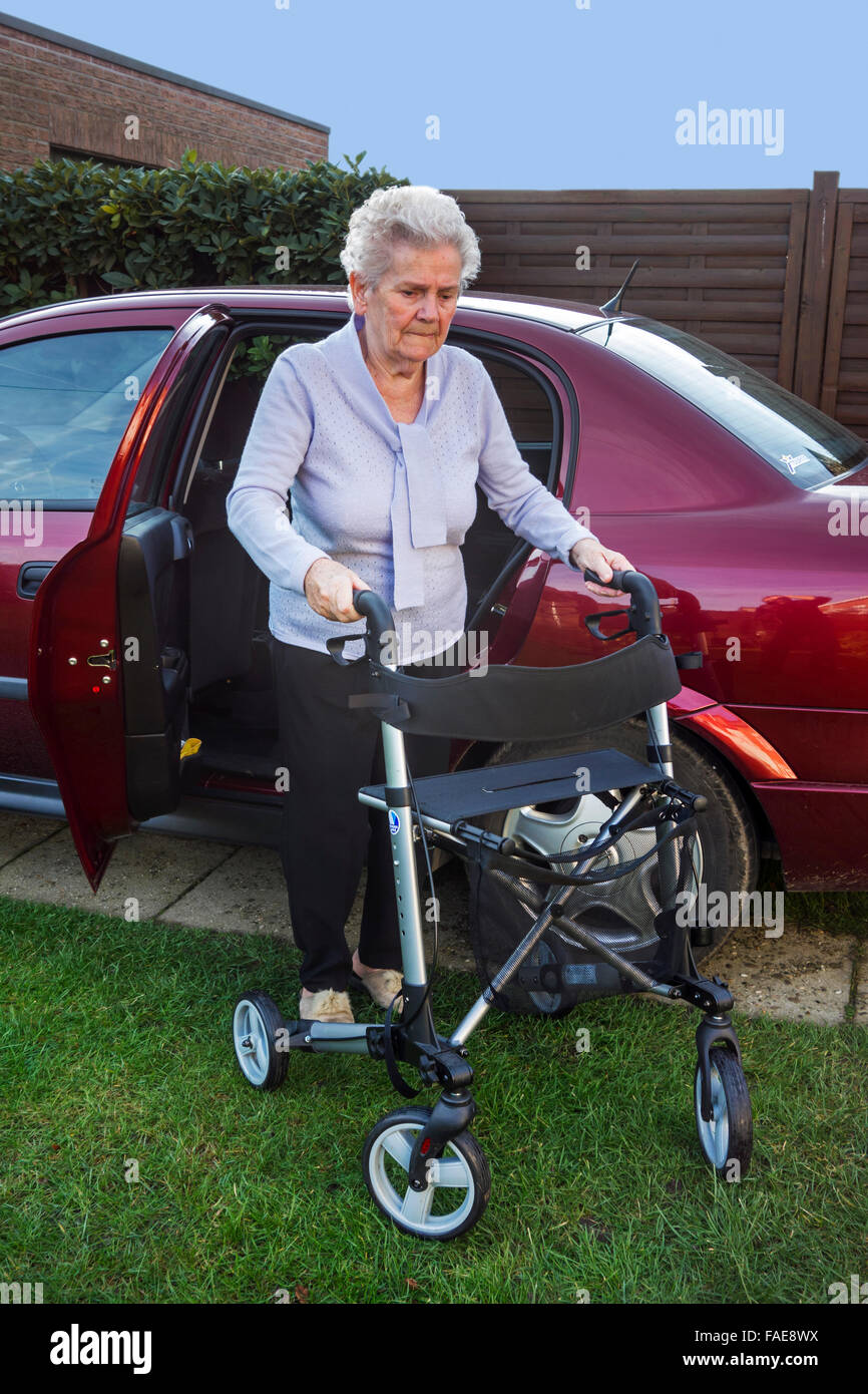 Donna anziana con rollator ruote / walker uscendo di auto sulla strada di casa Foto Stock