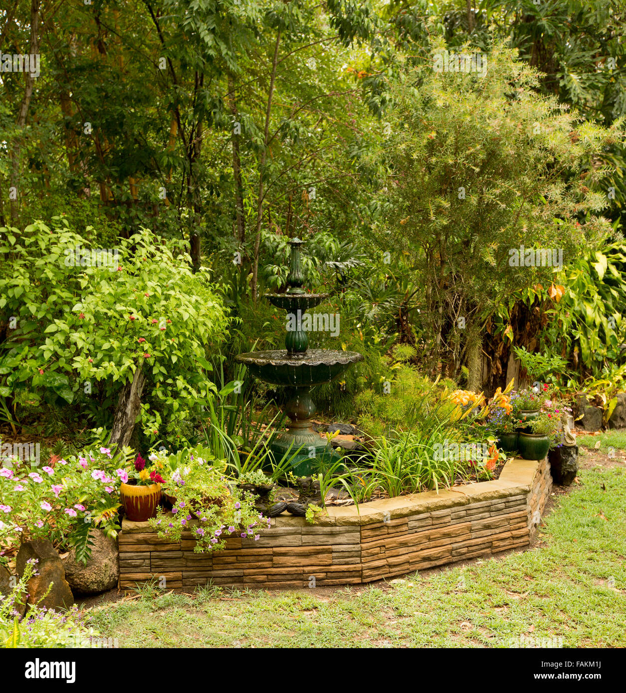 Un paesaggistico giardino con fontana ornata / acqua caratteristica circondata da muro di pietra, densi arbusti, alberi, fiori colorati e prato in Australia Foto Stock