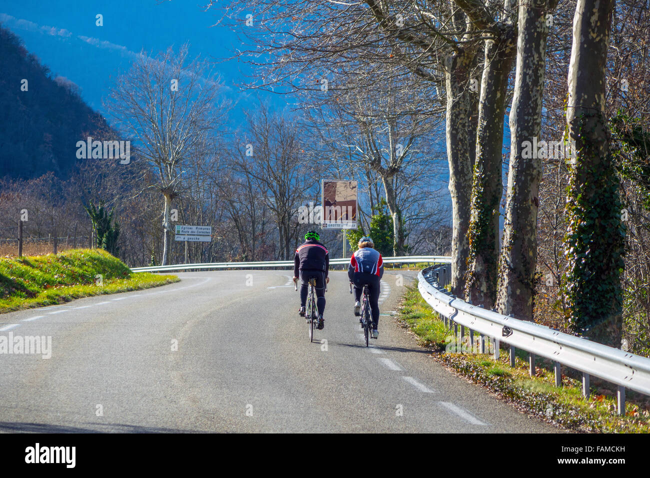 Stock Photo - Due adulti maschi cycle lontano dalla telecamera lungo una strada francese con montagne in lontananza Foto Stock