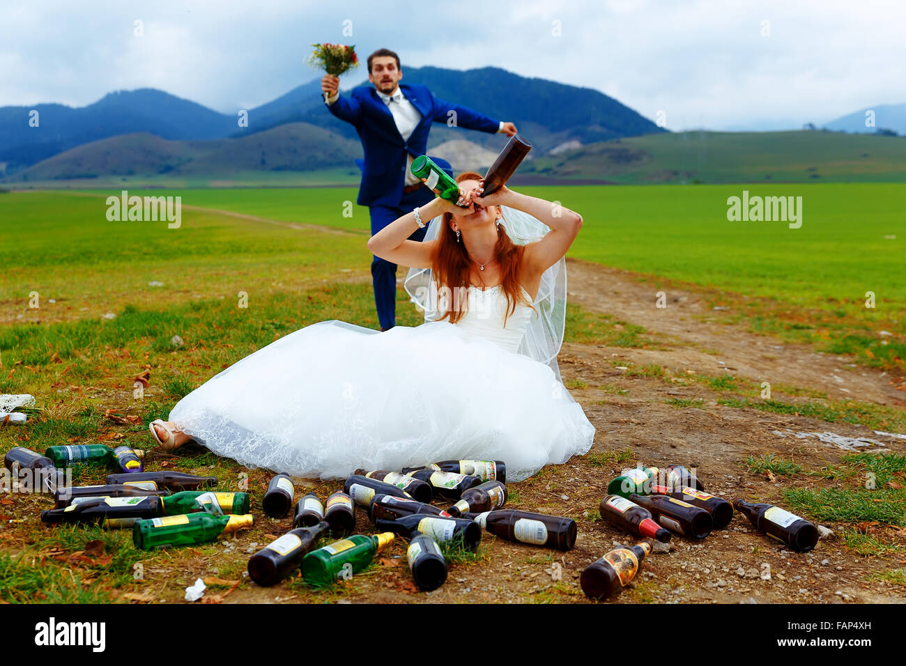 Drunken sposa con un sacco di svuotare le bottiglie di birra nel paesaggio di montagna e lo sposo racing a lei- strano concetto di matrimonio Foto Stock