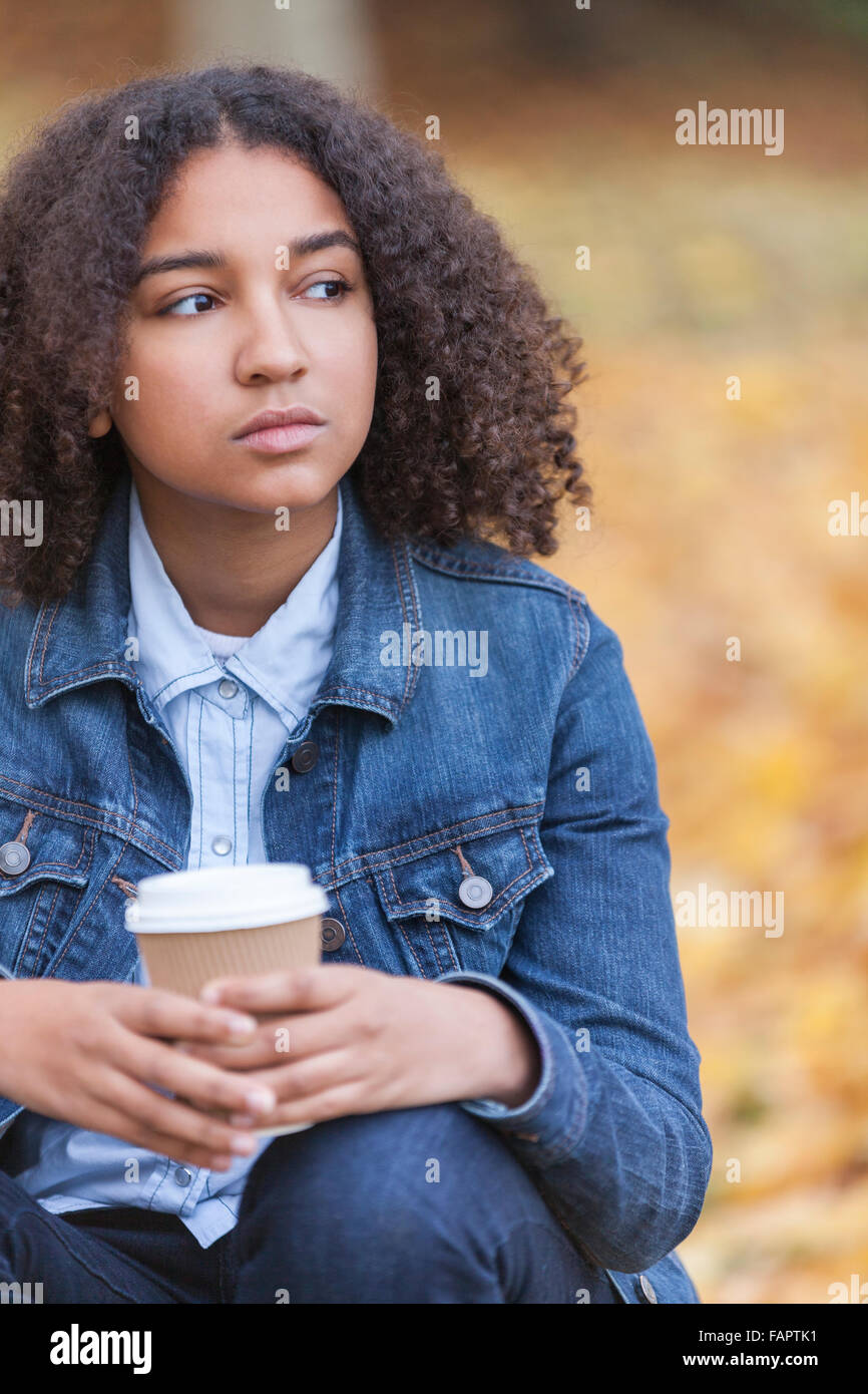 Bellissima gara di misto americano africano ragazza adolescente femmina giovane donna a bere il caffè da asporto al di fuori seduta in un parco Foto Stock