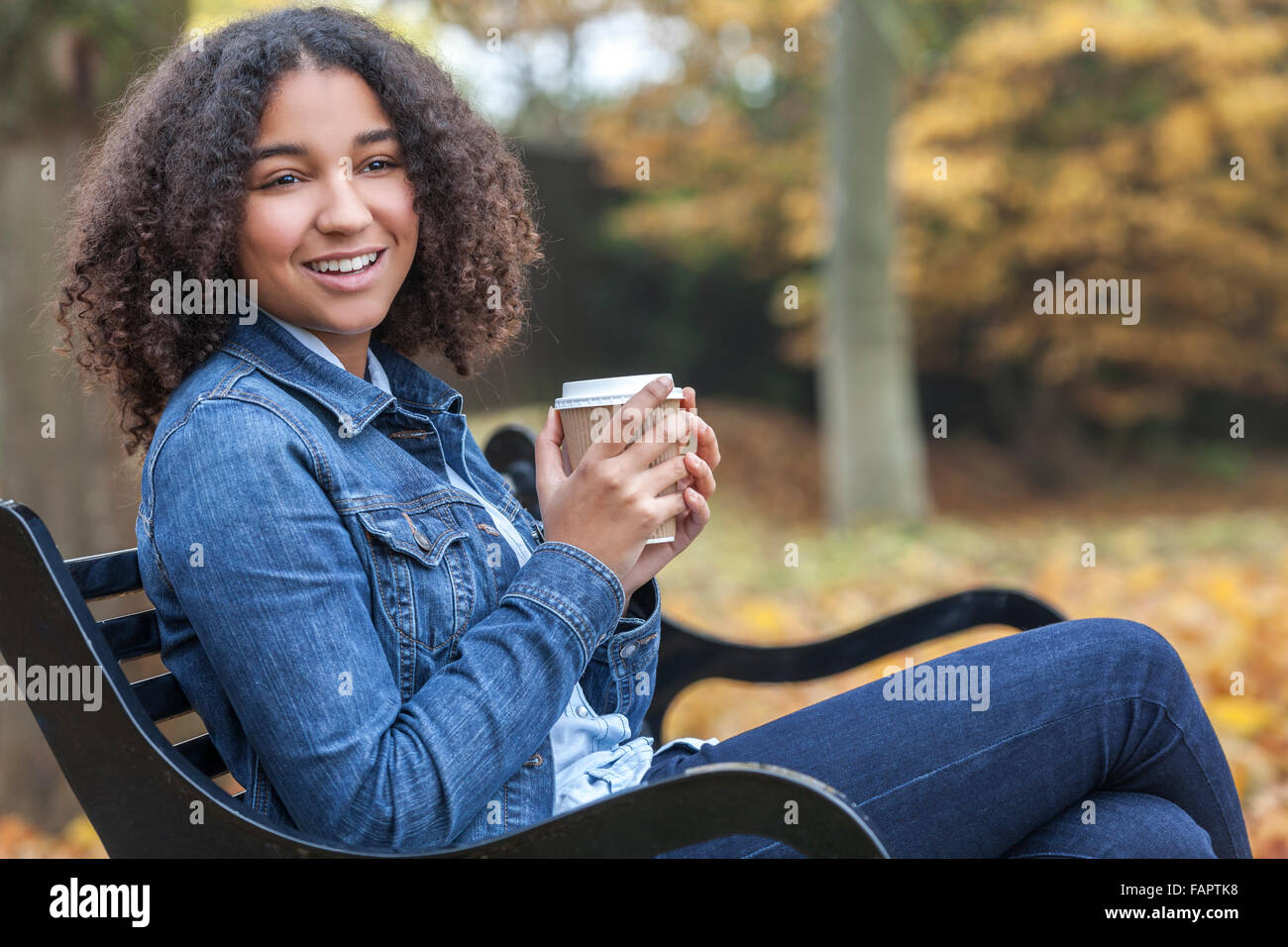 Bella felice razza mista americano africano ragazza adolescente femmine giovane donna sorridente di bere il caffè al di fuori seduta sul banco di lavoro Foto Stock