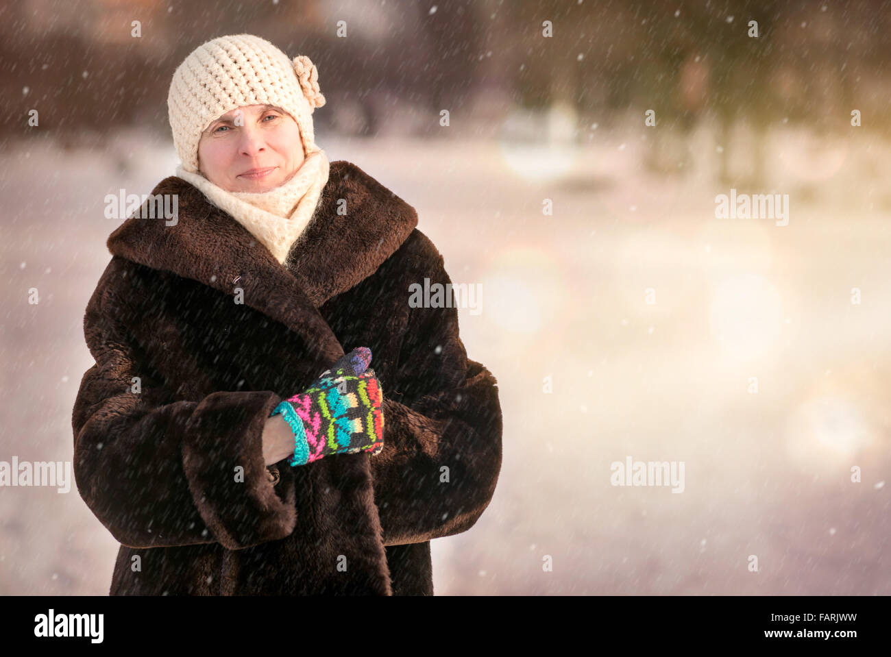 Un inverno ritratto di un sorridente senior adulto donna che indossa un cappello di lana, una sciarpa e guanti colorati, con un background di neve Foto Stock