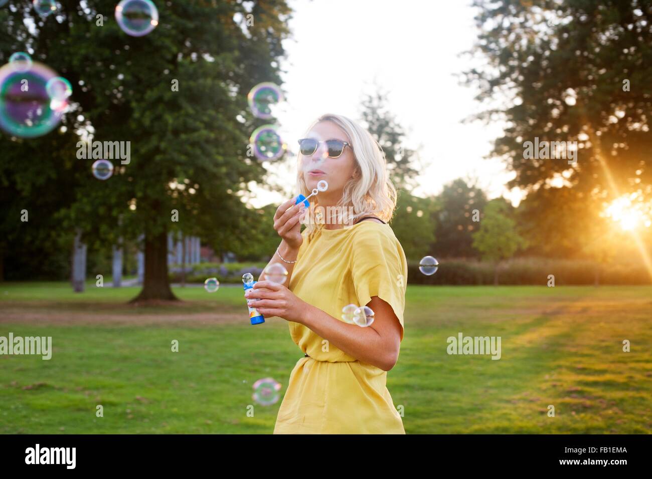 Ritratto di giovane donna che indossa abiti di colore giallo a soffiare bolle in parco al tramonto Foto Stock