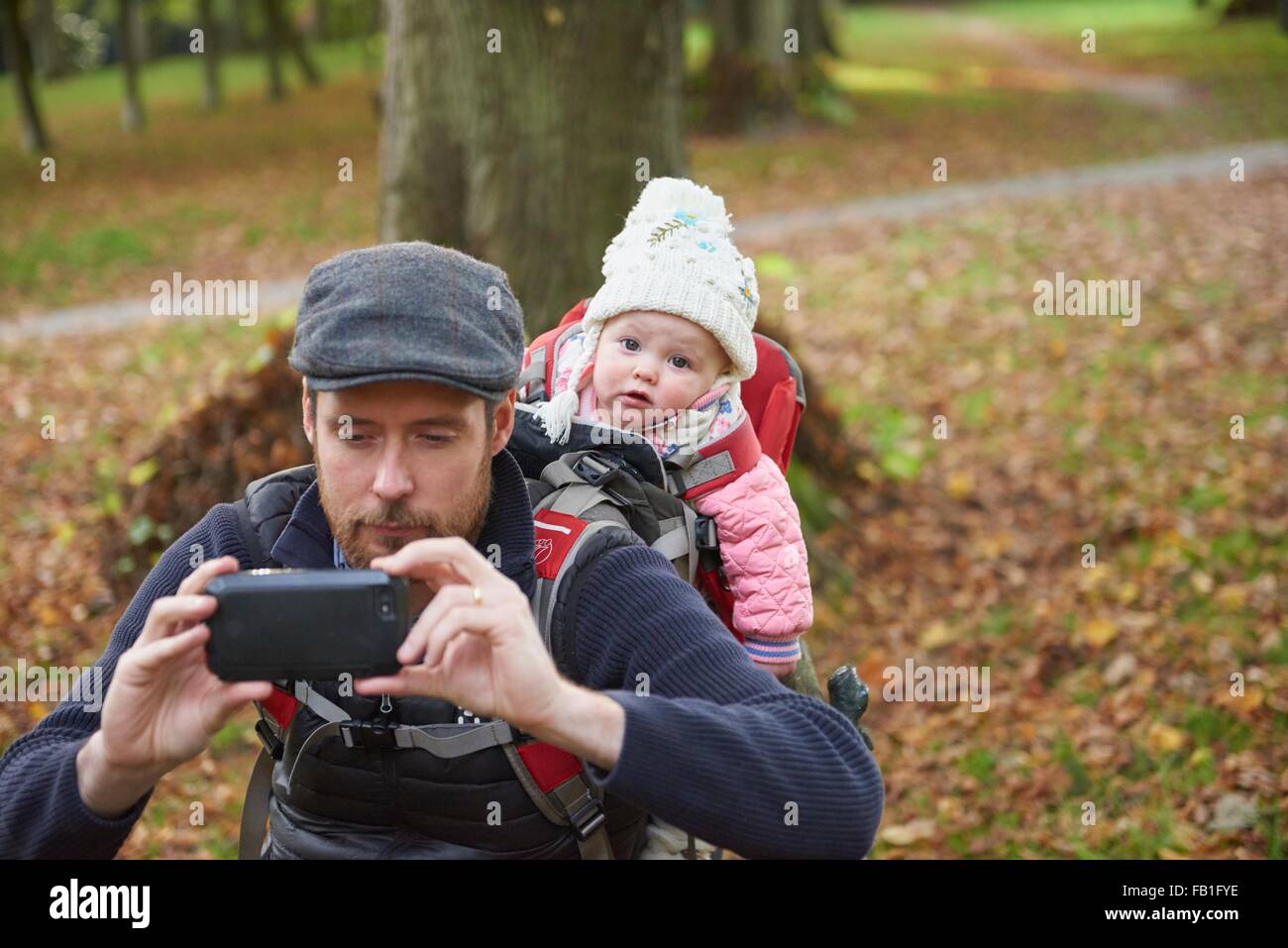 Metà uomo adulto in park che indossa il tappo piatto portante figlia sul retro in baby carrier tenendo selfie utilizza lo smartphone Foto Stock
