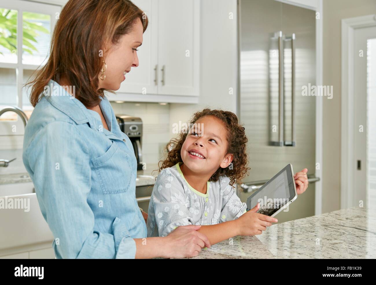 Madre e figlia al bancone cucina utilizzando digitale compressa, cercando di sorridere Foto Stock