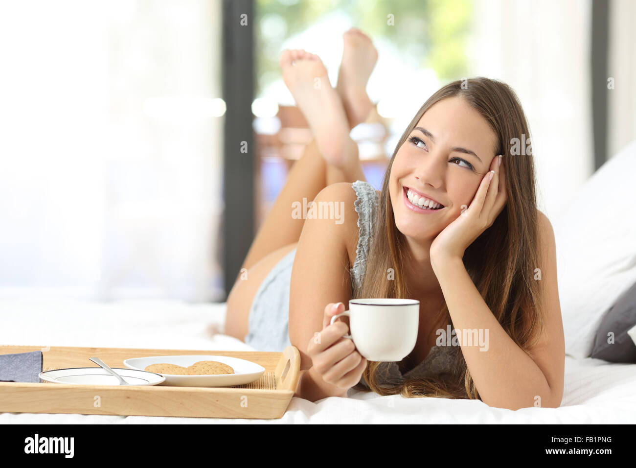 Felice ragazza avente la colazione tenendo una tazza di caffè giacente sul letto e guardando lateralmente Foto Stock